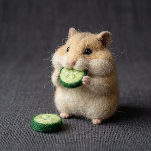 May include: A needle-felted hamster figurine crafted from tan and cream wool fibers. The hamster is holding a small, round, green felted cucumber slice to its mouth, while a second identical cucumber slice rests on the surface nearby.
