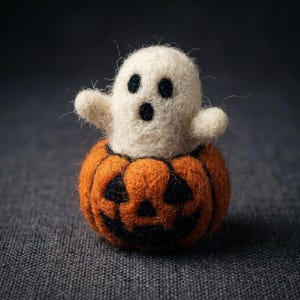 Può includere: Una figura di fantasma in lana infeltrita color crema con due occhi neri e una piccola bocca nera, che emerge dalla parte superiore di una zucca rotonda in lana infeltrita arancione con un volto di jack-o'-lantern nero e occhi triangolari.