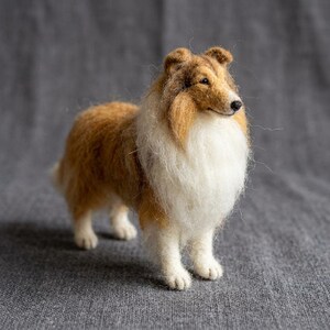 May include: A needle-felted figurine of a Rough Collie dog. The sculpture features a textured coat with a mix of golden-brown and white wool fibers. The dog is standing in a profile position, showcasing its pointed ears, long snout, and black nose.