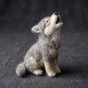 May include: A needle-felted wool sculpture of a small wolf pup sitting and howling with its head tilted upward. The figure features a textured coat of mixed gray, white, and tan fibers, with a black nose and an open mouth showing a dark interior.