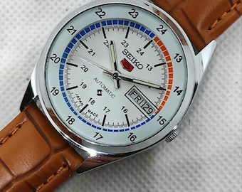 May include: A Seiko 5 automatic wristwatch with a white dial, silver-tone hands, and a day-date window displaying FRI 29. The dial features a 24-hour scale with blue and orange accents. The watch is attached to a brown leather strap with a textured finish.