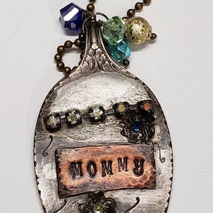 May include: A silver spoon necklace with a copper tag that says "MOMMY". The spoon is decorated with rhinestones, beads, and flowers.