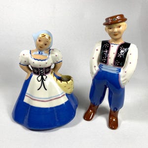 May include: Two ceramic figurines depicting a man and woman in traditional attire. The woman wears a blue and white dress with a bonnet and holds a basket. The man is in blue pants, a white shirt, and a black vest with floral accents.