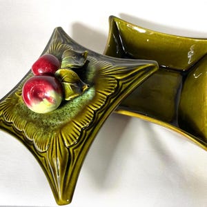 May include: Two olive green ceramic dishes with a starburst design. One dish features red and yellow ceramic fruit and leaves. The dishes have a glossy finish and are set against a white background. The dishes are decorative.