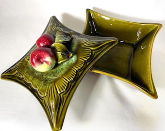 May include: Two olive green ceramic dishes with a starburst design. One dish features red and yellow ceramic fruit and leaves. The dishes have a glossy finish and are set against a white background. The dishes are decorative.
