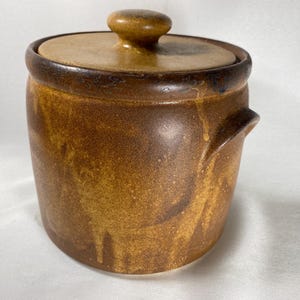 May include: A ceramic jar with a lid, featuring a warm brown and tan color scheme. The jar has a cylindrical shape with a slightly flared rim and a small handle. The lid has a rounded knob. The overall design suggests a rustic or vintage aesthetic.