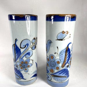 May include: Two ceramic shot glasses with a white base, adorned with blue floral and leaf patterns, and a brown rim. One glass features a bird and butterfly design. The glasses are approximately 5 inches tall.