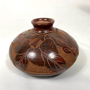 May include: A small, round, brown ceramic vase with a carved design of leaves and fruit. The vase has a slightly flared lip and a smooth, matte finish.
