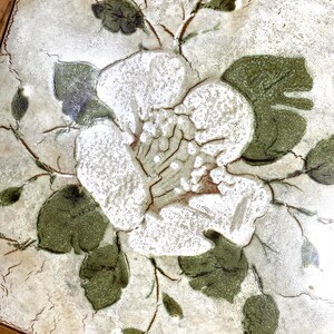 May include: A white ceramic tile with a floral design. The tile features a white flower with green leaves and a brown wooden frame.