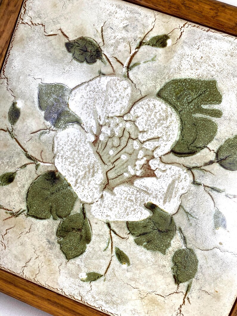 May include: A white ceramic tile with a floral design. The tile features a white flower with green leaves and a brown wooden frame.