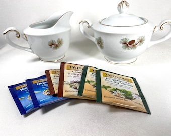 May include: A white porcelain creamer and sugar bowl set with gold trim and a pinecone design. Several tea packets are in front of the set, including flavors like Night, Camomile Honey, and Peppermint & Creamy Vanilla.