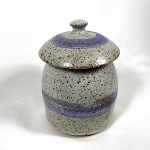 May include: A speckled ceramic jar with a lid. The jar is cylindrical with a light gray base and a wide lavender band. The lid is rounded with a matching lavender stripe and a small knob. The jar is likely handmade.