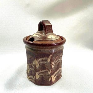 May include: A small, brown and white ceramic jar with a lid and a handle. The jar has a swirling pattern and is perfect for storing sugar, spices, or other small items.