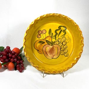 May include: A yellow ceramic pie plate with a scalloped edge, featuring a hand-painted design of fruit, including apples, grapes, and cherries. The plate is displayed on a metal stand, with a bunch of grapes and oranges to the left.