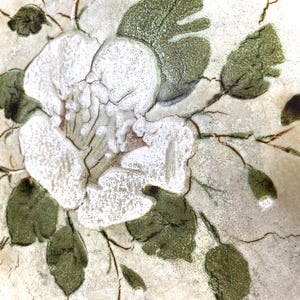 May include: A white ceramic tile with a hand-painted design of a white flower with green leaves. The tile has a textured surface and a rustic look.
