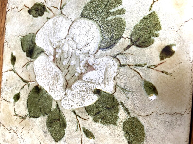 May include: A white ceramic tile with a hand-painted design of a white flower with green leaves. The tile has a textured surface and a rustic look.