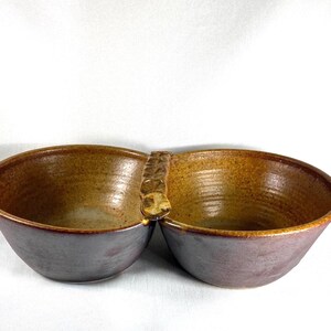 May include: A handmade ceramic serving bowl with two connected compartments. The interior is a warm brown, while the exterior has a muted purple hue. The handle is textured, adding a unique touch to this functional piece.