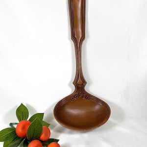 May include: A decorative wooden ladle with a long handle and an oval bowl. The ladle is a medium brown color and features ornate carvings along the handle and bowl. The handle has a heart-shaped design at the top. A sprig of orange fruit and green leaves is in the lower left.