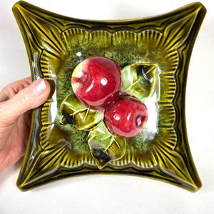 May include: A square, ceramic dish with a raised design of two red apples and green leaves. The dish has a glossy, olive green glaze with a scalloped edge. The apples are a vibrant red with a glossy finish.