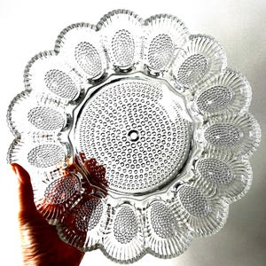 May include: Clear glass egg serving platter with a scalloped edge. The platter features oval-shaped indentations for holding eggs, with a textured, dotted pattern. The center has a circular design with small holes. The rim has a decorative, ridged pattern.