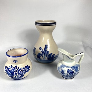 May include: Three small ceramic vases in a blue and white Delftware style. The largest vase has a windmill scene, the smallest vase has a sailboat scene, and the middle vase has a floral design.