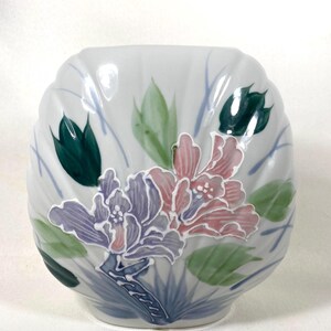 May include: A white ceramic vase with a scalloped design. The vase is decorated with hand-painted flowers in shades of purple, pink, and green. The floral design is set against a backdrop of blue and green leaves.