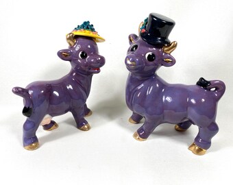 May include: Two glossy purple ceramic cow figurines standing side by side. One cow wears a yellow hat with blue floral accents, while the other wears a black top hat with small red and blue details. Both figures feature gold-painted horns and hooves.