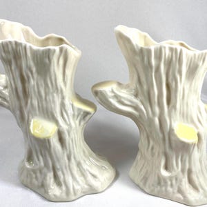 May include: Two off-white ceramic vases shaped like tree stumps with textured bark details and small yellow accents on the sides. Each vase features an irregular, open top rim and a wide, flared base, mimicking the appearance of a natural wood log.