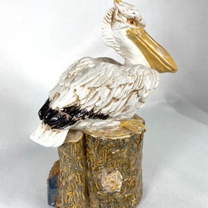 May include: A ceramic pelican figurine perched on a tree stump. The pelican is primarily white with black wing accents and a yellow beak. The tree stump is brown with a textured, bark-like appearance. The figurine is a decorative item.