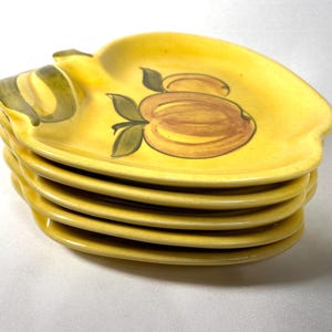 May include: A stack of six yellow ceramic apple-shaped plates. Each plate features a hand-painted design of a brown apple with green leaves. The plates have a curved edge and a small handle.