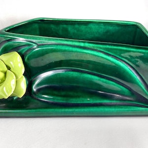 May include: A glossy, emerald green ceramic planter with a stylized, light green flower accent. The rectangular planter has a curved design and a deep interior, suitable for holding plants or other decorative items.