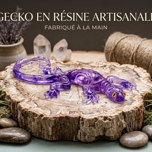Può includere: Una statuetta di geco in resina viola traslucida con dettagliati motivi scheletrici e occhi arancioni brillanti. Il testo GECKO EN RÉSINE ARTISANALE FABRIQUÉ À LA MAIN è visualizzato sopra l'oggetto, appoggiato su una fetta di legno.