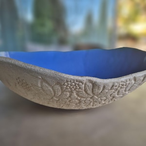 Può includere: Una ciotola in ceramica poco profonda e allungata con un interno smaltato blu vibrante. L'esterno presenta una superficie beige ruvida e non smaltata con un dettagliato motivo floreale e a foglie in rilievo che copre tutto il lato esterno.