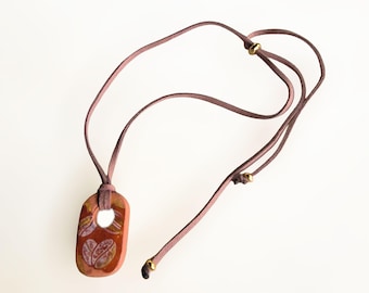 May include: A necklace featuring a terracotta-coloured oval pendant with a central hole and white leaf-like patterns. The pendant hangs from a mauve-coloured cord that includes two small gold-toned metal beads for adjustable length.