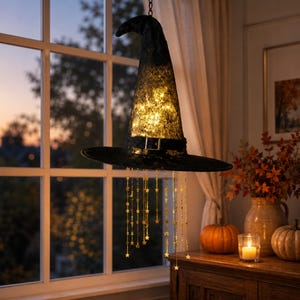 May include: A black witch hat decoration featuring a textured lace overlay and a central buckle. The interior is illuminated by warm white string lights, and gold star-shaped charms hang from the brim on thin metallic chains for display.