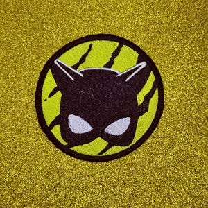 May include: A circular embroidered patch featuring a black cat mask silhouette with white eyes centered on a lime green background. The design includes black claw mark accents radiating from the mask, all enclosed within a thick black circular border.