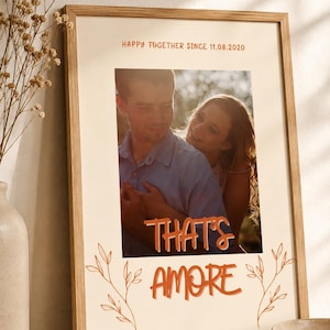 Könnte beinhalten: Ein gerahmter vertikaler Druck mit einem Foto eines sich umarmenden Paares. Der Text HAPPY TOGETHER SINCE 11.08.2020 steht oben und THAT'S AMORE in orangefarbenen Buchstaben unten, flankiert von einfachen, liniengezeichneten botanischen Motiven.