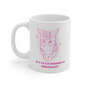 Könnte beinhalten: Ein weißer Keramikbecher mit einer rosa Strichzeichnung einer mürrisch aussehenden Katze, die von kleinen Sternakzenten umgeben ist. Unter der Katze steht der Text "MY 9 TO 5 IS RUINING MY PERSONALITY" in einer fetten, rosa serifenlosen Schrift.