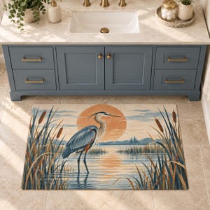 Peut inclure: Un tapis de bain rectangulaire orné d'une illustration imprimée représentant un héron bleu debout dans une eau peu profonde, entouré de grandes massettes. L'arrière-plan montre un grand soleil orange se couchant sur un lac calme.
