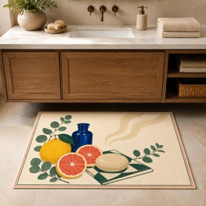 May include: A rectangular bath mat featuring a cream-colored background with a graphic illustration of a yellow citrus fruit, two grapefruit slices, a dark blue glass bottle, a beige bar of soap, and green leafy branches with steam swirls.