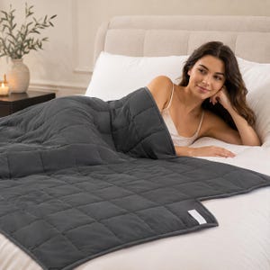 Puede incluir: Una manta pesada de color gris carbón oscuro con un patrón de costuras en cuadrícula en toda la superficie. La manta tiene una pequeña etiqueta de tela rectangular de color blanco sujeta en la esquina inferior.