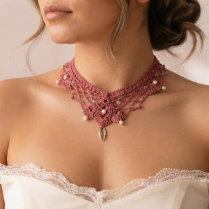 Op de afbeelding: Een oudroze gehaakte choker-halsketting met ingewikkeld draadwerk, kleine witte bolvormige kralen verwerkt in het patroon en een enkele metalen druppelvormige hanger die in het midden van het decoratieve kantontwerp hangt.