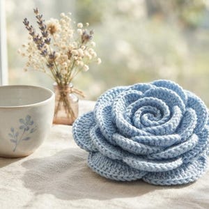 May include: A light blue crocheted rose with layered, textured petals arranged in a spiral pattern. Beside the flower sits a ceramic mug featuring a small blue floral design and a glass bottle containing a bouquet of dried lavender and white flowers.