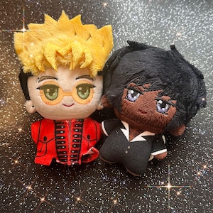 May include: Two plush dolls with embroidered facial features. The left doll has spiky yellow hair, round orange glasses, and a red jacket. The right doll has dark brown skin, black hair, and wears a black suit with a white collar and buttons.