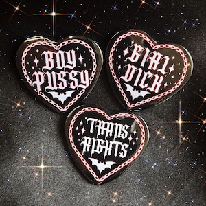 Peut inclure: Trois badges noirs en forme de cœur avec une écriture gothique blanche et des bordures en chaîne rouge. Les badges portent le texte BOY PUSSY, GIRL DICK et TRANS RIGHTS, chacun orné d'une petite chauve-souris blanche et d'étoiles scintillantes.