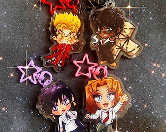 May include: Four acrylic keychains featuring chibi-style anime characters. Each charm has a star-shaped clasp in red, black, pink, or purple. The characters include a blonde figure in a red coat, a dark-haired figure in a suit, a dark-haired figure in a white shirt, and an orange-haired figure in a white shirt and tie.