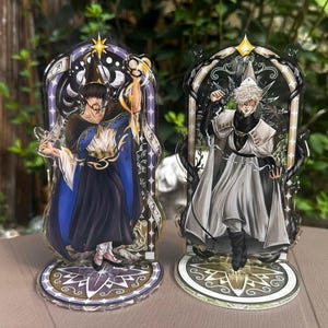May include: Two acrylic character standees featuring illustrated figures in arched frames. The left figure wears a dark robe with a blue cape and pointed hat. The right figure wears a white and black outfit with a pointed hat. Both have circular bases.