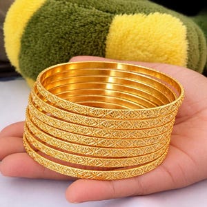 Op de afbeelding: Een stapel van acht goudkleurige metalen armbanden. Elke armband is voorzien van een herhalend geometrisch sterpatroon dat in het buitenoppervlak is gegraveerd. De armbanden hebben een gepolijste, reflecterende afwerking en zijn cirkelvormig.