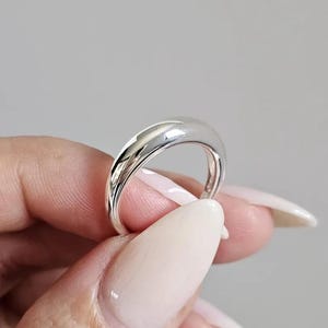 以下が含まれることがあります： 滑らかで丸みを帯びた、わずかに先細りになった銀色の磨き上げられた指輪。金属の表面は光をよく反射し、明るいハイライトと、円形の全体にわたって一定の厚みを保つすっきりとした曲線的なシルエットが特徴です。
