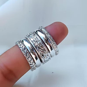 May include: A set of five silver-toned rings stacked together. The collection includes two smooth, polished bands, one band with a detailed leaf-like pattern, one band featuring an intricate floral design, and one band with a textured geometric pattern.